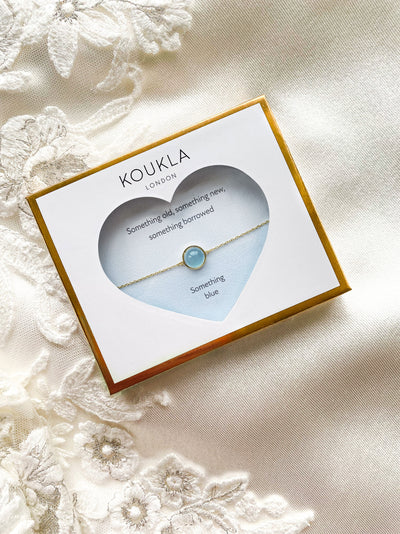 Dainty gold bridal bracelet with blue aquamarine stone in giftable wedding packaging, laid on wedding dress