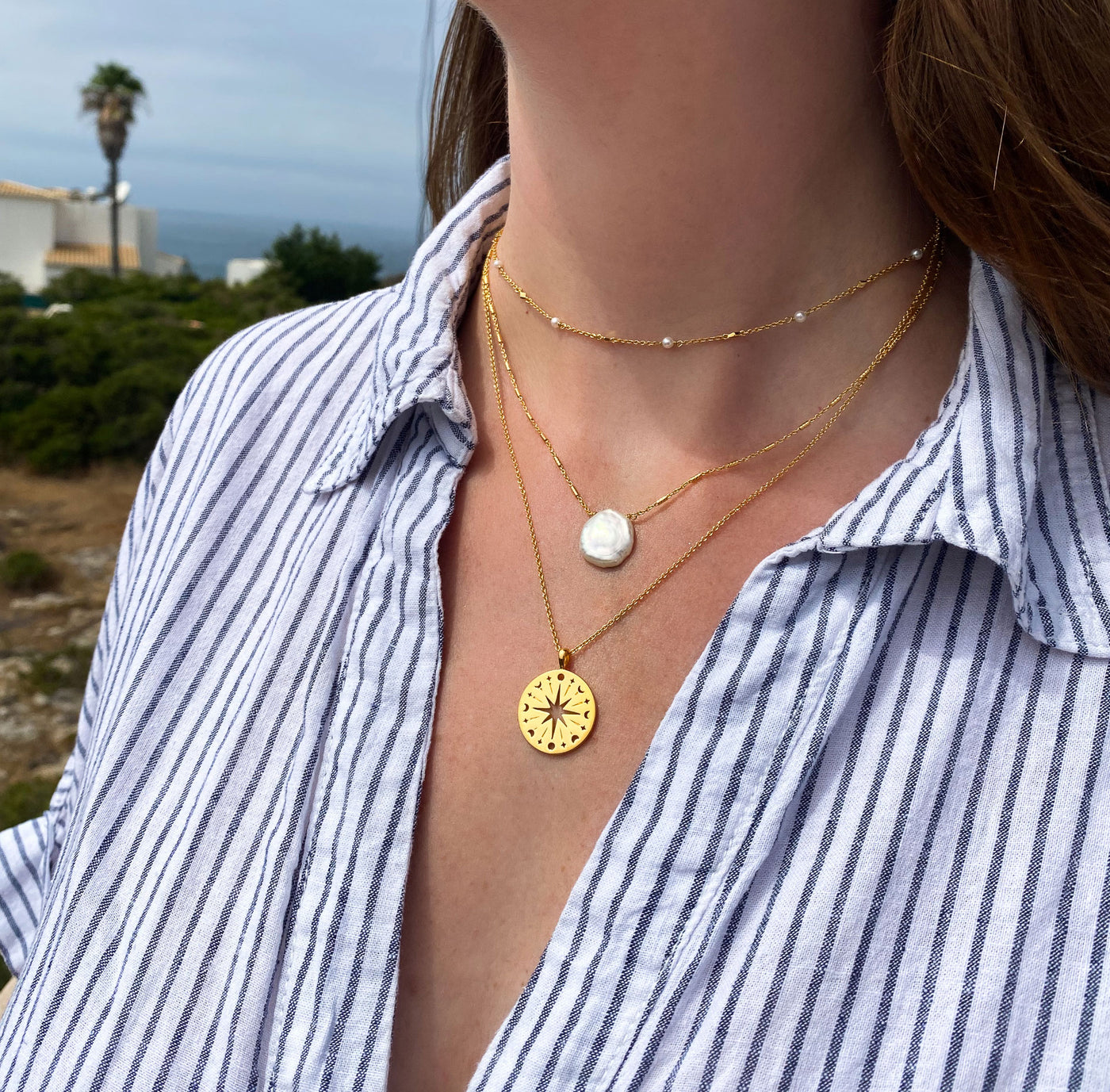 Person wearing layered gold coin compass necklace, freshwater pearl necklace and pearl choker with a Greek scenic background
