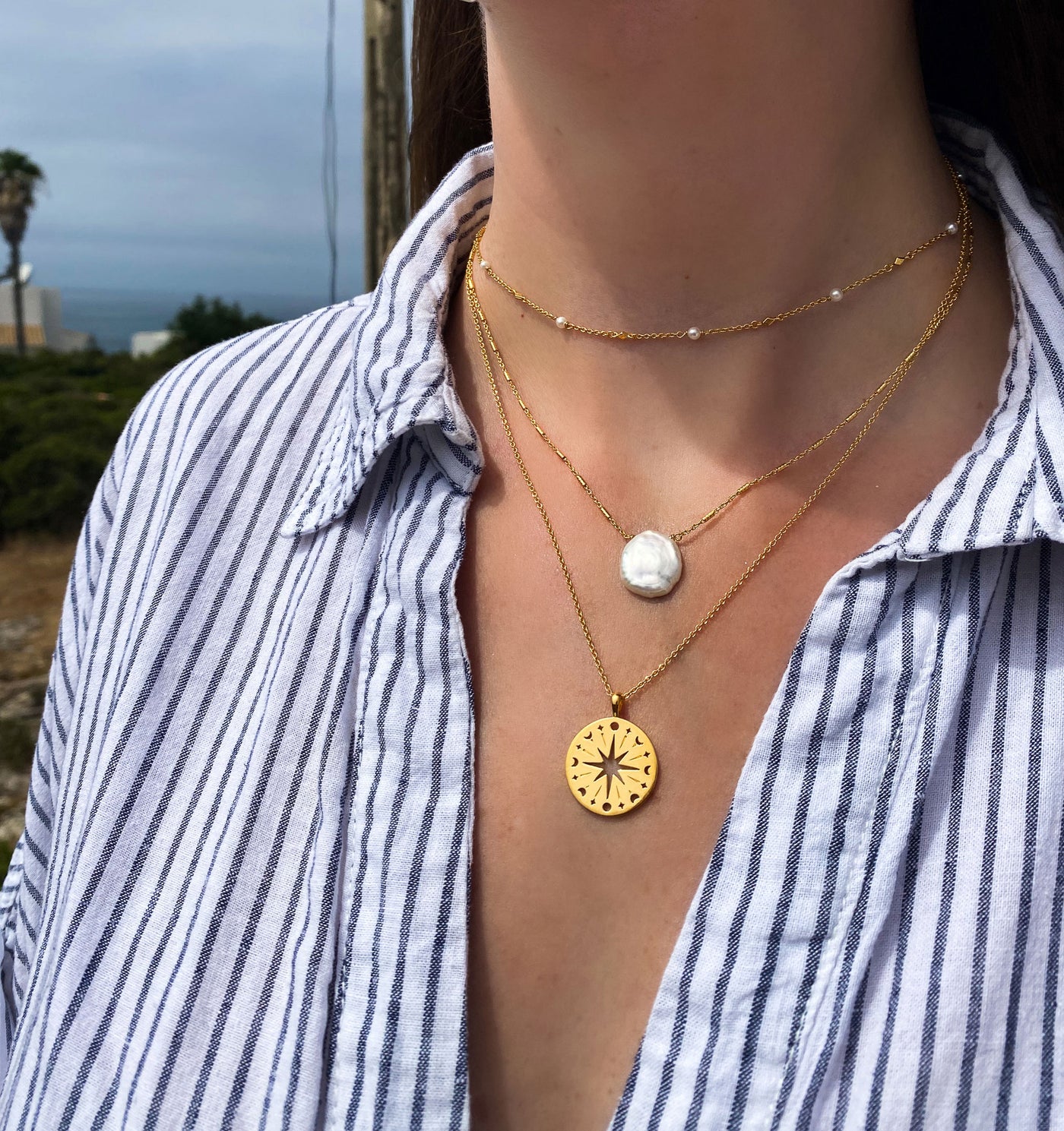 Person wearing layered gold coin and freshwater pearl necklaces and pearl choker with a Greek scenic background