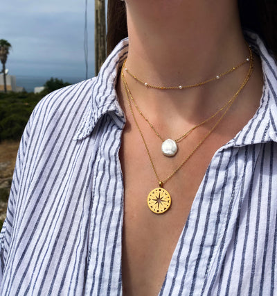 Person wearing layered gold coin and freshwater pearl necklaces and pearl choker with a Greek scenic background