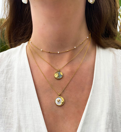 Close-up of a person wearing an amazonite gold sun necklace, a mother of pearl moon pendant layered with a dainty pearl choker, set against a blurred natural background.