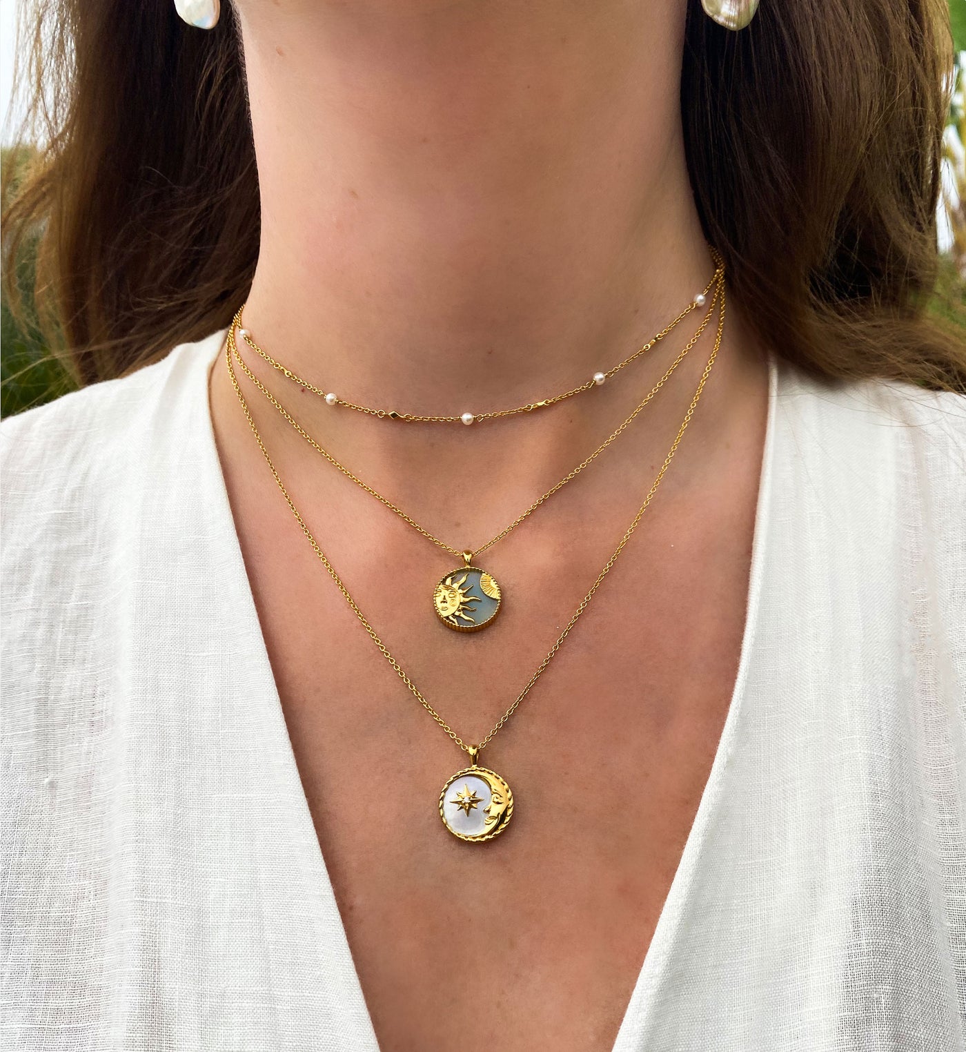 Close-up of a person wearing an amazonite gold sun necklace, a mother of pearl moon pendant layered with a dainty pearl choker, set against a blurred natural background.